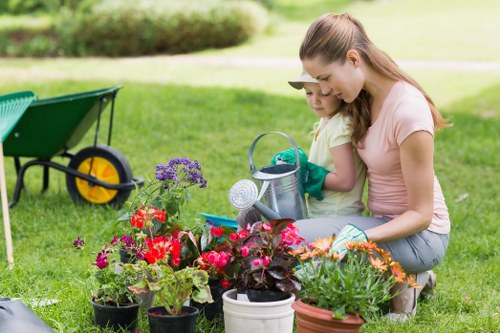 A woman and a young girl are crouching on a well-maintained, lush green lawn in a garden, engaging in gardening activities. The woman is smiling and holding a small garden trowel, while the girl is wearing gardening gloves and looking attentively at the plants. Surrounding them are various potted flowering plants displaying vibrant reds, purples, and oranges, positioned in the foreground. In the background, there is a wooden wheelbarrow with a yellow wheel, partially visible, and a paved patio area adjacent to a building, with a neat hedge and trees further in the distance. The garden features a mix of cultivated flower beds and a neatly cut lawn, with natural daylight illuminating the scene under a clear sky, indicating pleasant weather. This outdoor space forms part of a typical residential garden in Camden, designed for gardening and outdoor leisure, aligning with services offered by Hedge Trimming Camden for maintaining pristine landscaped gardens.