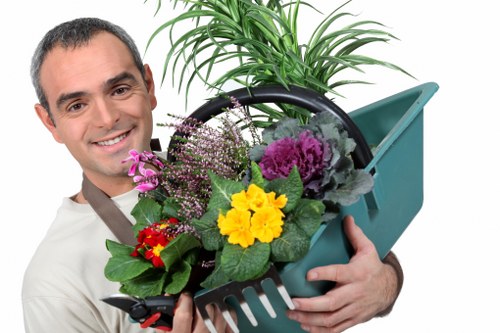 A smiling man holding a tool basket filled with potted plants, including vibrant yellow, purple, and red flowers, and green foliage, standing outdoors in a garden setting. Behind him, there is a well-maintained lawn with dense, healthy grass, bordered by a variety of shrubs and small trees, indicating a landscaped yard typical of Camdenâ€™s residential areas. The environment appears bright with natural daylight, suggesting clear weather. The scene highlights outdoor gardening activities, suitable for gardening and landscaping services offered by Hedge Trimming Camden, with visible details of garden elements like soil, paving, and plant containers contributing to the suburban garden aesthetic frequently found in the Camden area.