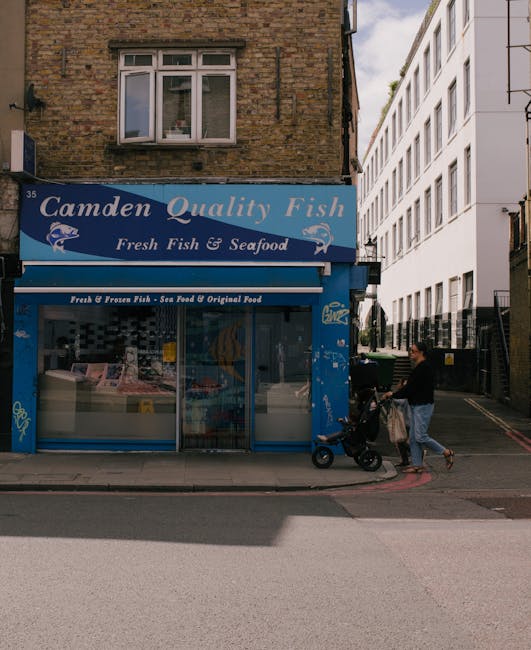 A view of a small urban shop named 'Camden Quality Fish' with a blue sign displaying 'Fresh Fish & Seafood' above a large glass window showcasing fish and seafood products. The shop is situated on a s