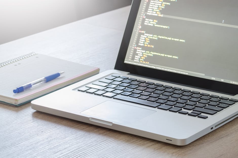 A silver laptop with a black keyboard displaying a code editor on the screen, placed on a light wooden desk. Next to the laptop, there is an open spiral notebook with a blue pen resting on top. The ba