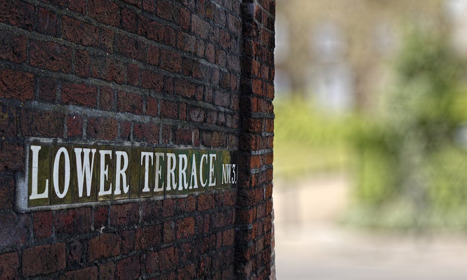 A close-up image of a brick wall with a street sign reading 'LOWER TERRACE NW3' attached to its surface. The bricks are dark red with a slightly rough texture, interspersed with black mortar. The sign